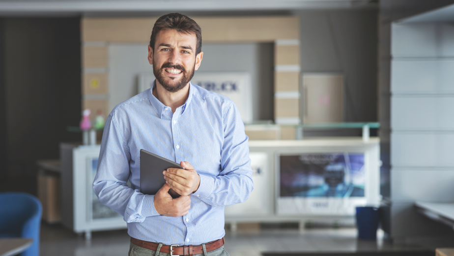 Smiling bearded businessman standing in lobby of shipping firm and holding tablet while looking at camera.